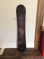 Photo of a full-length Gnu men's snowboard standing upright against a white wall, showing the board's dark color and grapevine graphic design.