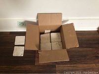 Box with approx 40 off white porcelain tiles stacked inside, and four tiles laid out on the floor around the box.