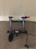 Photo showing blue Max Racer Pro Trainer indoor bike trainer with quick-release clamp, and black front wheel riser block on carpet.