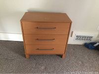 Front view showing three drawers and metal handles; item placed against wall on carpeted floor.