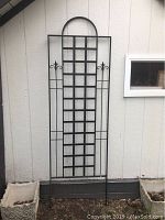 Front view of black metal garden trellis placed against light-colored wall near window and planters. Shows full height and structural details.