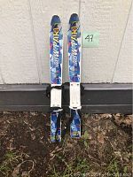 Blue toddler cross country skis with white bindings labeled 'Snow Park', photographed standing upright outdoors against a wall with a number 47 label.