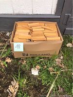 Full view of one box filled with parquet flooring wood pieces outdoors on grass by a building wall.