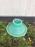 Green plastic Christmas tree base with four metal eyebolts sitting on grass near a building.