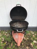 Open black charcoal BBQ grill showing the metal cooking grate and warming rack. Mounted on an orange metal wheeled stand. Shows signs of use and rust.