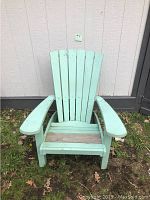 Photo of a large solid wood Adirondack chair painted light green with some paint wear and discoloration on the seat, placed outdoors against a building wall.