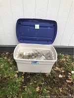 Large beige plastic tote with blue lid, filled with glass Mason jars, photographed outdoors on grass against siding wall.