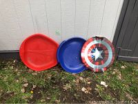Three kids sleds arranged outside against a white wall on grass and fallen leaves; includes red and blue plastic round saucer sleds and a Captain America-themed sled.