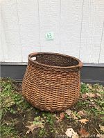 Wicker basket placed outdoors on grass and leaves with a white wall background. Shows basket shape, color, and weave detail clearly.