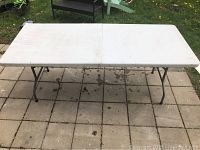 White rectangular collapsible table set up outdoors on tiled ground. The table top is white plastic with a fold line in the middle and folding black metal legs underneath.