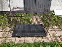 Full view of large rectangular metal dog crate placed outdoors on stone tiles with grass surrounding. Black wire frame with removable plastic tray at the bottom.