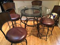 Round glass top table surrounded by four metal frame chairs with padded brown round seats and embossed metal backs showing wear.