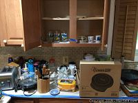 Overview of kitchen counter with multiple items including boxed NuWave Pro induction cooktop, mugs, knife block with knives, glasses, and kitchen utensils.
