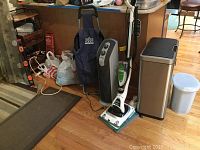 Overview showing the Whole Foods shopping cart, Holmes fan, Shark hard floor cleaner, and the two trash cans on wooden floor near the wooden cabinet.