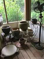 Overview of standing fan, plant stands, small table and various ceramic and metal planters arranged on a wooden floor with outdoor view