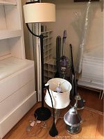 Wide view showing four lamps including tall black floor lamp with cream shade, white bucket lamp base, two silver pendant lamps, and a box with vacuum cleaner in background