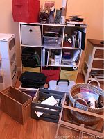 Photo showing the 9-cubby white storage unit filled with various storage containers, baskets, and art supplies including papers, brushes, paints, and spray bottles. Several wooden items and rolling storage cart are placed in front on the floor.