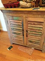 Full front view of the wooden cabinet showing the multi-colored distressed painted surface with slatted doors and two metal pull handles.