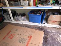 Shelf with three bins and one cardboard box containing various tools and hardware items in a garage or workshop space