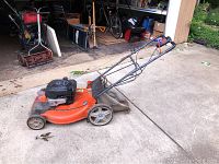Side and angled view of the orange Husqvarna lawn mower showing metal deck, engine, handle, and wheels on concrete surface in front of garage. Grass bag attached at rear.