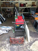 Wide view showing red reel mower with detached handle, folding table, gas cans, utility cart with supplies, luggage rack, garden chairs, and other garden tools.