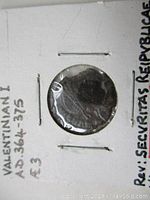 Front side of the coin inside a white cardboard holder showing the profile of Emperor Valentinian I with the date and denomination noted on the holder.