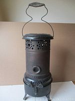Front view of the antique stove heater showing cylindrical body with ventilation holes and three supporting legs.