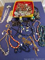 Full layout photo showing an assortment of vintage costume jewelry including bead necklaces, chains, and earrings arranged on a table with a yellow-bordered box holding many pieces.
