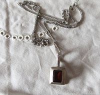 Pendant and chain arranged loosely on a light-colored embroidered fabric background, showing full length of chain and pendant shape and size.