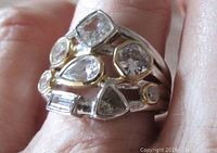 Close-up of the ring worn on a finger showing pear, trillion, baguette, round, and cushion shaped cubic zirconia stones with gold-plated highlights and polished finish.