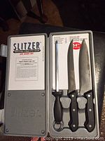 Top view of the Slitzer 3 piece knife set inside the grey plastic case showing two knives and sharpening steel along with product information card.