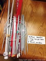 Photo shows a collection of knitting needles on a wooden surface with a handwritten note indicating the contents: 1 large hairpin lace, several sets, 2 round needles.