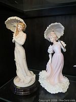 Photo showing two porcelain lady figurines holding parasols, one on wooden stand.