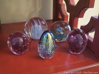 Five crystal glass eggs photographed on a red surface near wooden furniture, showing different colors and internal designs.