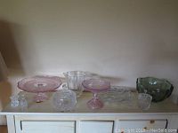 Wide view of all glass and crystal items arranged on a white cabinet including pink Gorham cake platter, candy dish, clear glass bud vase, green bowl, bell, salad servers, covered dish, and salt and pepper shakers