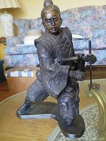Full view of the seated samurai figurine showing overall pose and armor details on a glass table with a couch in the background.