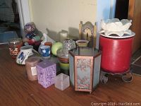 Photo shows a collection of assorted candles, candle holders, and decorative items arranged on a wooden surface, including a large red candle with white lotus flower top, purple and brown pillar candles, various glass and metal candle holders.