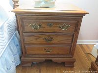 Front view of one wooden night stand showing three drawers with ornate brass handles and natural wood finish.