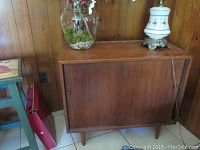 Full view of wooden mid-century modern cabinet with sliding doors and tapered legs, placed against wood-paneled wall.