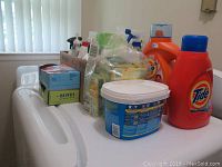 Overview of cleaning supply bottles including Tide orange detergent and plastic containers on a white base under window blinds