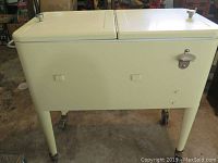 Front view of the cream-colored stand-up cooler showing dual lids, metal bottle opener fixed on front, legs with caster wheels.