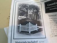 Black and white assembly and instruction manual with image of mini corner picket fence installed outdoors on lawn near garden area under tree