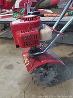 Close-up of engine and tines showing red engine housing and metal soil tilling blades.