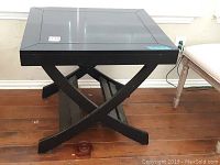 Black lacquer wood square side table with crossed legs and visible glossy finish on wood surface, sitting on wooden floor.