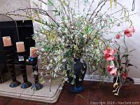Black ceramic urn with plastic greenery and terra cotta pot with silk orchid placed nearby, shown next to candle holders for height reference.