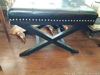 Black rectangular padded footstool with button tufting and silver nailhead trim on black wooden X-shaped base.
