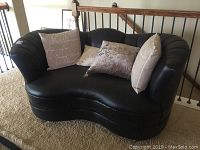 Photo showing front angle of black leather retro style love seat with 4 pillows placed on it, on carpet flooring near stair rail.