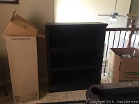 Front view of the black bookcase showing three open shelves, with some packed boxes around it.