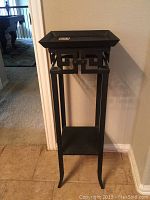 Full view of black metal plant stand showing geometric Greek key style design and two shelves.