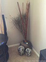 Photo showing two oriental style ceramic jars with black floral patterns and pointed lids on beige carpet floor next to a large textured ceramic pot filled with dried decorative branches and grasses, placed in corner of room.